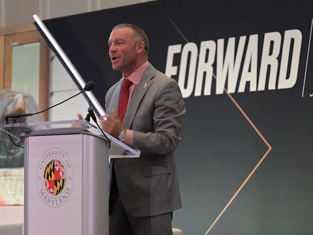 UMD vice president Jim Harris, gesturing as he speaks from behind a lectern. A black background with the words "Forward: The University of Maryland Campaign for the Fearless" is behind him.